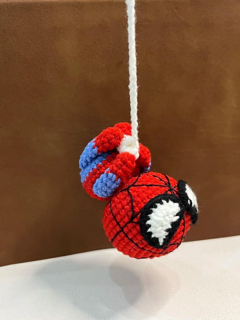 May include: A crocheted amigurumi figure of a superhero character with a round red head, black web-like stitching, and large white eyes. The figure is suspended by a white yarn string and features red and blue crocheted limbs attached to the top.