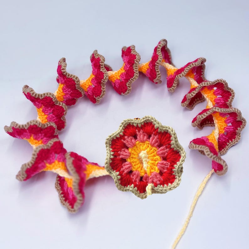 May include: A handmade crochet floral garland featuring vibrant red, pink, and yellow flowers with brown edging. The garland is arranged in a spiral, with a central flower. The craftwork is displayed on a white surface.