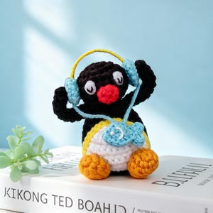 May include: A handmade crochet penguin figurine with a black body, white belly, and yellow and orange accents. The penguin wears blue headphones and a blue fish-shaped pendant. The figurine is approximately 4 inches tall.