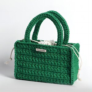 May include: A handmade, emerald green crochet tote bag with two arched handles. The bag features a textured stitch pattern and a white drawstring closure. A small metal tag with the word "handmade" is attached to the front.