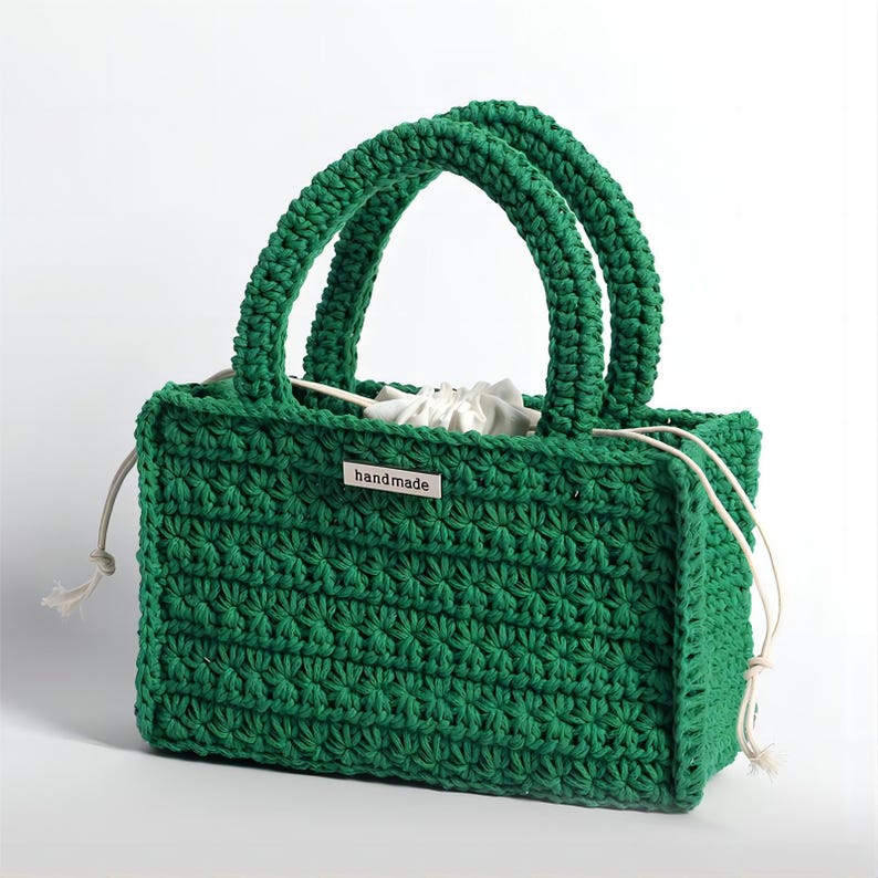 May include: A handmade, emerald green crochet tote bag with two arched handles. The bag features a textured stitch pattern and a white drawstring closure. A small metal tag with the word "handmade" is attached to the front.