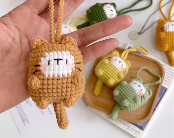 Op de afbeelding: Een set van vijf gehaakte katten sleutelhangers in bruin, groen en geel. Elke sleutelhanger heeft een kleine kat met een wit gezicht en een lange staart.