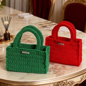 May include: Two handmade crochet tote bags. One is emerald green, the other is bright red. Each bag has a rectangular shape with a rounded handle and a small metal tag that reads "handmade" or "Elliemofs". The bags sit on a marble-patterned surface.