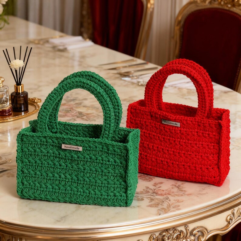 May include: Two handmade crochet tote bags. One is emerald green, the other is bright red. Each bag has a rectangular shape with a rounded handle and a small metal tag that reads "handmade" or "Elliemofs". The bags sit on a marble-patterned surface.