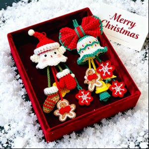May include: A red velvet box containing crocheted Christmas decorations. The ornaments include a Santa Claus, a bell, snowflakes, gingerbread men, and mittens. A card with "Merry Christmas" is also in the box.