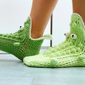 Puede incluir: Pantuflas de ganchillo verdes con forma de cocodrilo con ojos blancos y un ribete blanco alrededor de la parte superior de la pantufla.