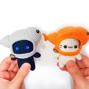 May include: Two crocheted plush toys, one with a gray and black color scheme and the other with a white and orange color scheme. Both toys are wearing crocheted hats.
