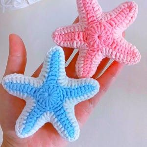May include: Two crocheted starfish in shades of pink and blue. The blue starfish has a white border, while the pink one is solid. The handmade items are held in a hand, showcasing the texture and detail of the yarn.