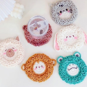 Op de afbeelding: Zes gehaakte onderzetters met dierengezichten. De onderzetters zijn in verschillende kleuren, waaronder wit, grijs, bruin, oranje en groen. Elke onderzetter heeft een ander dierengezicht, waaronder een kat, een konijn, een lama, een schaap, een kikker en een beer.