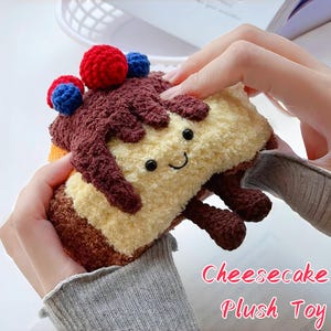 Puede incluir: Un peluche con forma de porción de tarta de queso, con una base amarilla y crema, glaseado marrón y bayas de ganchillo rojas y azules. El juguete tiene una cara sonriente y pequeñas patas marrones. El texto "Cheesecake Plush Toy" es visible.