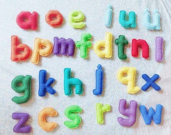 May include: A selection of brightly coloured, crocheted lowercase letters displayed on a white surface. The letters are in various colours, including red, orange, yellow, green, blue, and purple. The letters have a soft, textured appearance.
