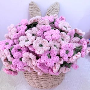 May include: A handmade crochet floral arrangement in a basket, featuring pink and light pink flowers with green centers. The basket is beige, and two beige bunny ears are attached to the back of the arrangement.