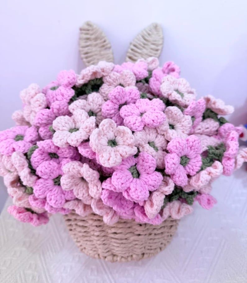 May include: A handmade crochet floral arrangement in a basket, featuring pink and light pink flowers with green centers. The basket is beige, and two beige bunny ears are attached to the back of the arrangement.