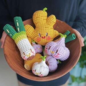 May include: A wooden bowl filled with crocheted amigurumi vegetables. The vegetables include a yellow ginger, a green onion, a purple garlic, and a white garlic.