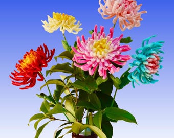 May include: A vibrant bouquet of handmade flowers in a dark vase against a blue gradient backdrop. The flowers are red, yellow, pink, and blue, with green leaves. The vase is dark brown.