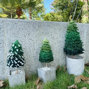 Könnte beinhalten: Drei dekorative gehäkelte Weihnachtsbäume in verschiedenen Grüntönen, jeweils in einem grauen quadratischen oder zylindrischen Topf gepflanzt. Die Bäume stehen auf Gras mit einer Steinmauer im Hintergrund.