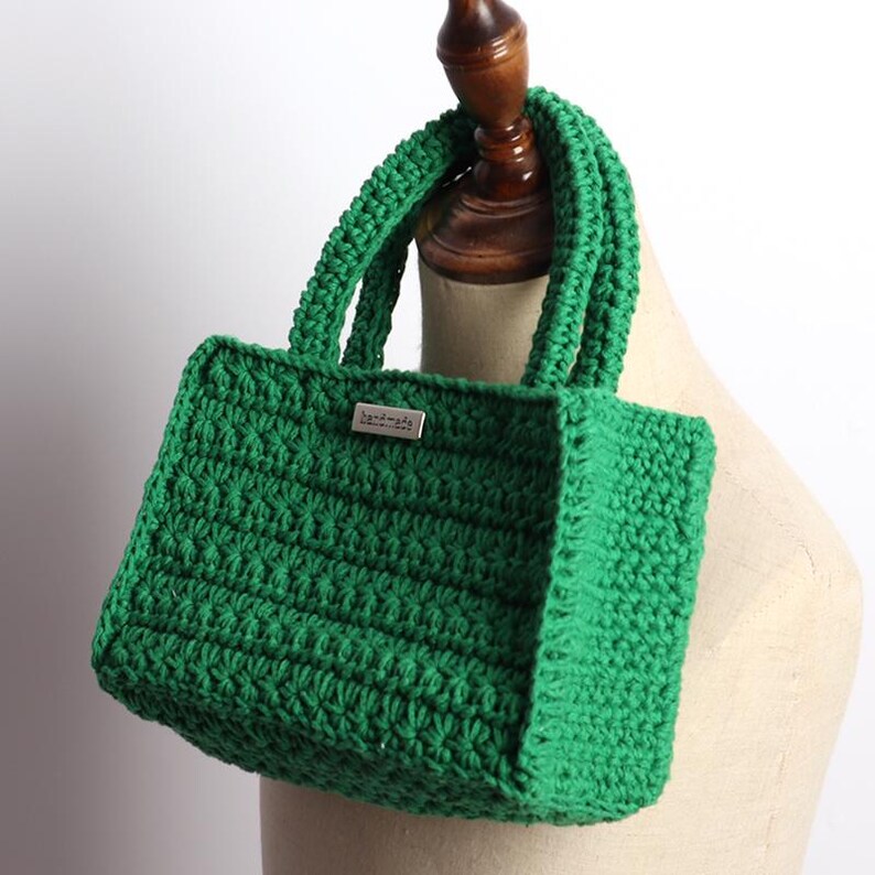 May include: A handmade, emerald green crochet tote bag. The bag features a rectangular shape with a textured stitch pattern and sturdy handles. A small, rectangular metal tag with the word "handmade" is attached to the front.