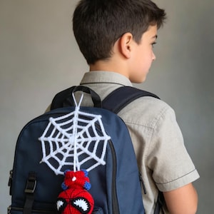 May include: A navy blue backpack featuring a crocheted accessory attached to the top handle. The accessory consists of a white yarn spiderweb hanging above a round, red and blue crocheted character head with large white eyes and black outlines.