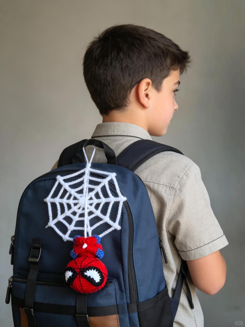 May include: A navy blue backpack featuring a crocheted accessory attached to the top handle. The accessory consists of a white yarn spiderweb hanging above a round, red and blue crocheted character head with large white eyes and black outlines.