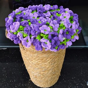 May include: A handcrafted crochet floral arrangement in a woven basket. The arrangement features purple and lavender flowers with green accents. The basket is made of natural-colored rope.
