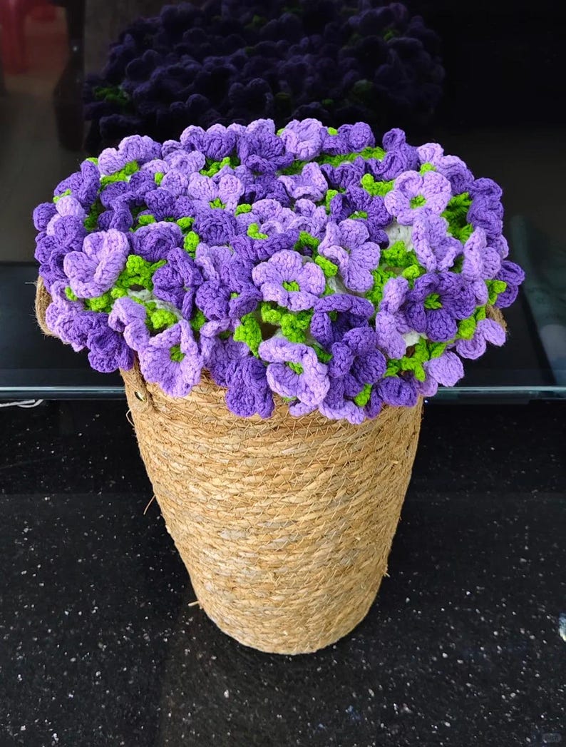 May include: A handcrafted crochet floral arrangement in a woven basket. The arrangement features purple and lavender flowers with green accents. The basket is made of natural-colored rope.