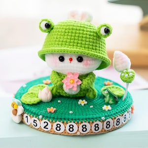 May include: A green crocheted frog with a white face and pink flower holding a green crocheted frog with a white face and pink flower. The frog is sitting on a green crocheted base with white numbers.