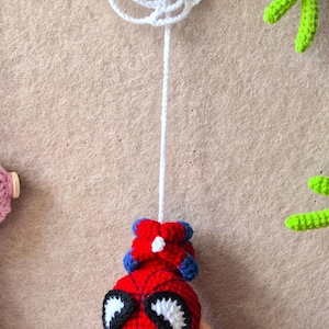 May include: A crocheted red and blue superhero character with large white eyes and black outlines, suspended by a long white yarn string from a white crocheted spider web. The character features a round head and small blue and red limbs.