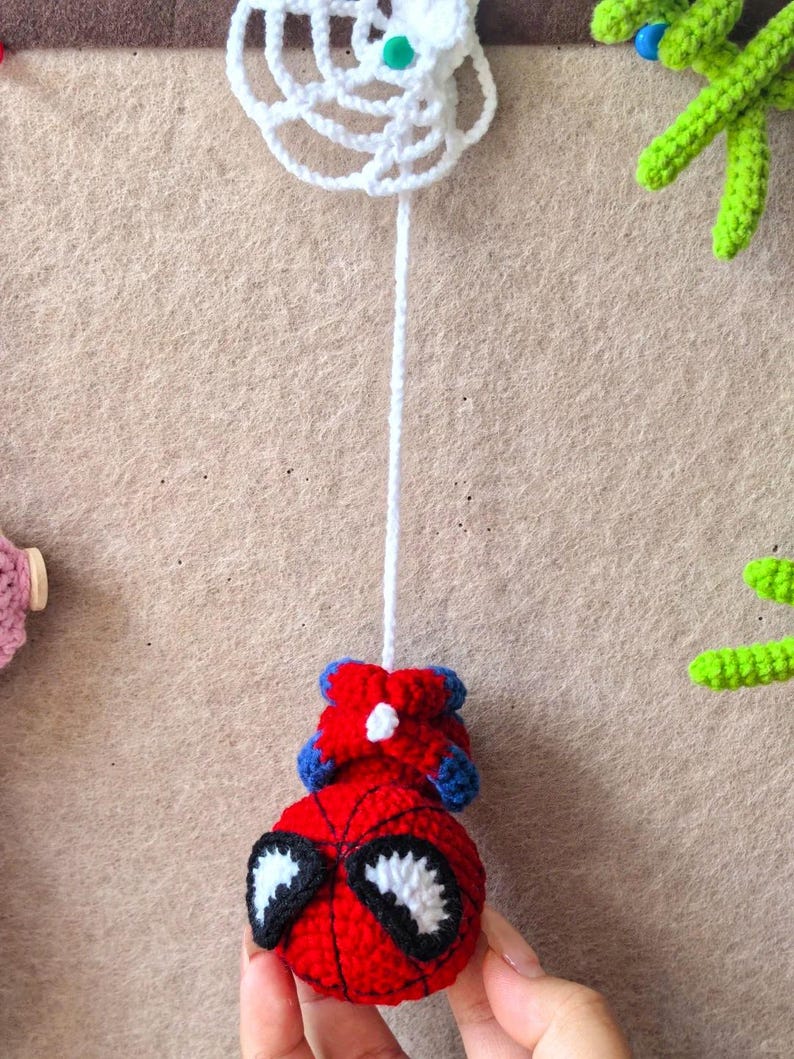 May include: A crocheted red and blue superhero character with large white eyes and black outlines, suspended by a long white yarn string from a white crocheted spider web. The character features a round head and small blue and red limbs.