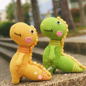 May include: Two crocheted dinosaur toys, one yellow and one green, with embroidered eyes and pink cheeks. The yellow dinosaur is standing on its hind legs, while the green dinosaur is sitting with its tail curled around its legs.