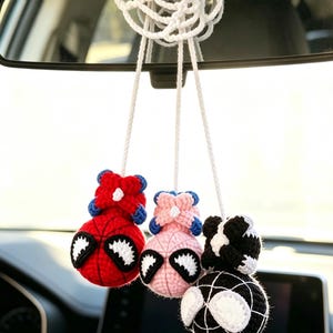 May include: A hanging car accessory featuring three crocheted spherical figures suspended by white cords from a white crocheted web-like base. The figures are red, light pink, and black, each with white eye patches and a small crocheted top element.