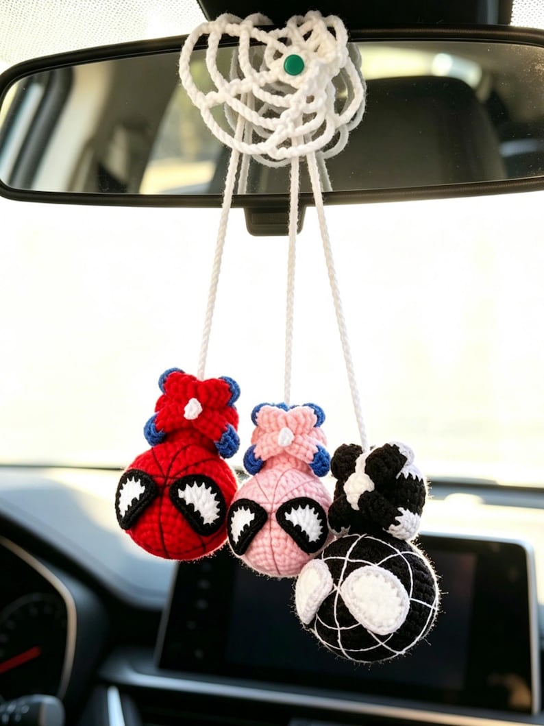 May include: A hanging car accessory featuring three crocheted spherical figures suspended by white cords from a white crocheted web-like base. The figures are red, light pink, and black, each with white eye patches and a small crocheted top element.