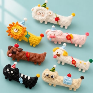 Op de afbeelding: Een verzameling van zeven gehaakte dierenfiguren, waaronder een leeuw, schaap, varken, hond en kat. Elk figuur is langwerpig, draagt een klein feesthoedje en heeft een uniek accessoire zoals een vis, appel of een patch met de tekst CUTE.
