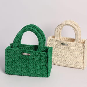 May include: Two handmade crochet tote bags. One is a vibrant green, the other is off-white. Both have a rectangular shape, sturdy handles, and a small metal tag that reads "handmade". The bags are displayed against a plain white background.