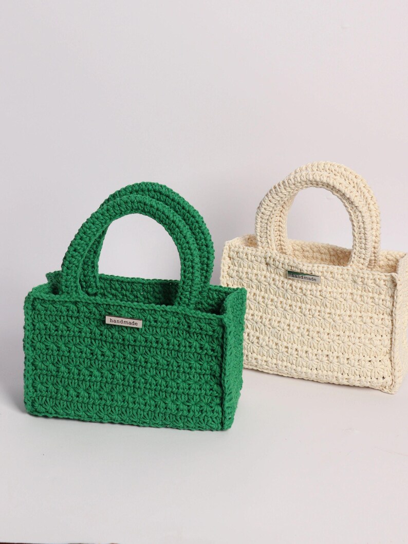 May include: Two handmade crochet tote bags. One is a vibrant green, the other is off-white. Both have a rectangular shape, sturdy handles, and a small metal tag that reads "handmade". The bags are displayed against a plain white background.
