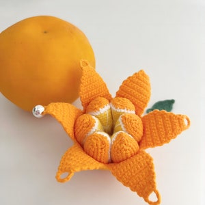 May include: A crocheted orange with a segmented design, resembling a peeled orange. The crocheted orange is made with orange yarn and has a white centre. The orange is sitting on a white surface.