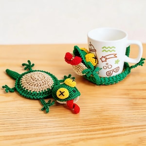Può includere: Sottobicchieri a forma di lucertola all'uncinetto fatti a mano in verde, beige, rosso e giallo. Un sottobicchiere è sotto una tazza bianca con illustrazioni a tema estivo. L'altro sottobicchiere è piatto. I sottobicchieri sono su una superficie di legno.