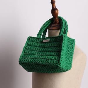 May include: A handmade, emerald green crochet tote bag with two handles. The bag has a rectangular shape and a textured stitch pattern. A small tag that says "handmade" is attached to the front of the bag.