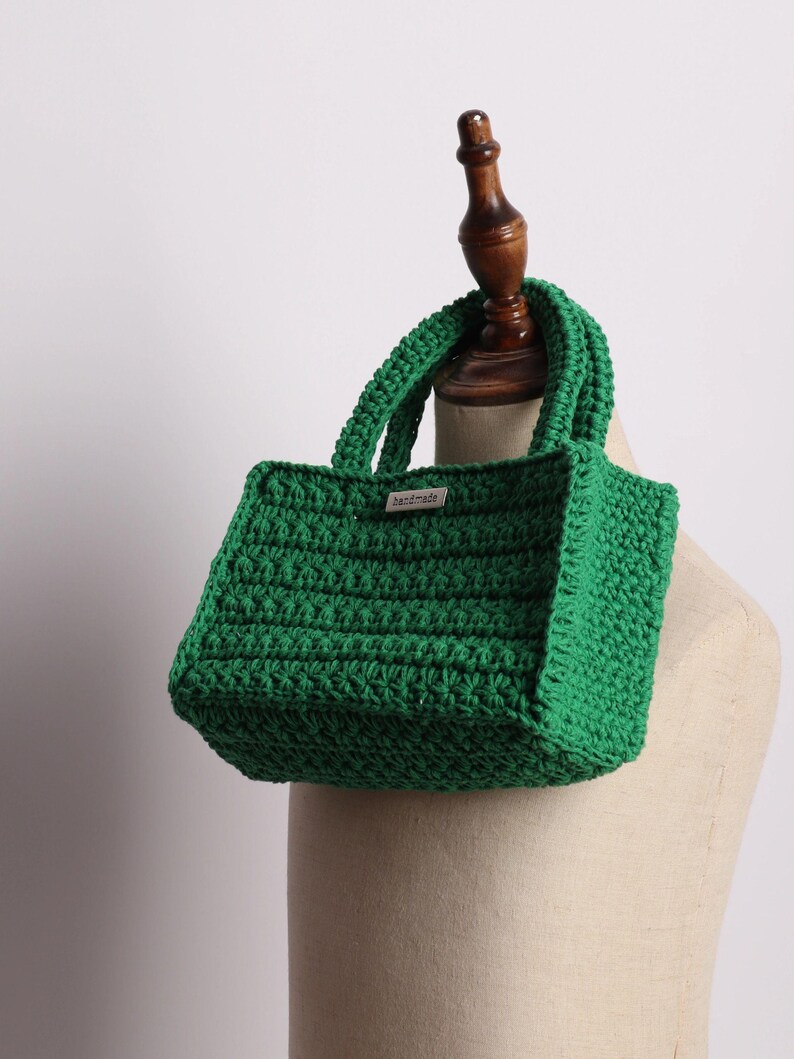 May include: A handmade, emerald green crochet tote bag with two handles. The bag has a rectangular shape and a textured stitch pattern. A small tag that says "handmade" is attached to the front of the bag.
