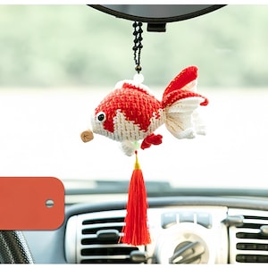 Puede incluir: Un adorno para coche de pez dorado tejido a ganchillo, rojo y blanco. El pez tiene una borla roja y un pequeño amuleto de madera. El adorno cuelga de una cadena de cuentas negras, delante del salpicadero de un coche.