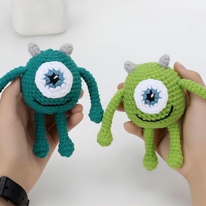 May include: Two crocheted green one-eyed monsters with gray horns and a smiling mouth. The monsters are green and teal.
