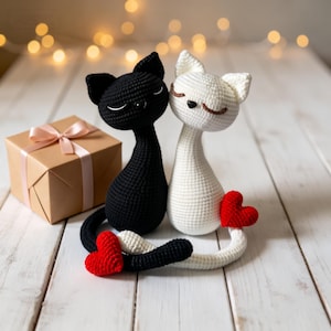 Könnte beinhalten: Gehäkelte schwarze und weiße Katzenfiguren mit roten Herzakzenten, die zusammen sitzen. Eine verpackte Geschenkbox mit rosa Schleife befindet sich in der Nähe. Die Katzen befinden sich auf einer weißen Holzoberfläche mit weichen Bokeh-Lichtern im Hintergrund.