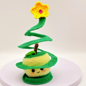 May include: A crocheted apple with a green stem and leaves, a yellow flower with a pink center, and a green spiral stem. The apple has a smiling face.