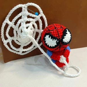 May include: A small, round, crocheted figure featuring a red and blue body with a large red head, black web-like stitching, and white eyes. The figure holds a long, white, crocheted spider web strand that connects to a circular white web piece.