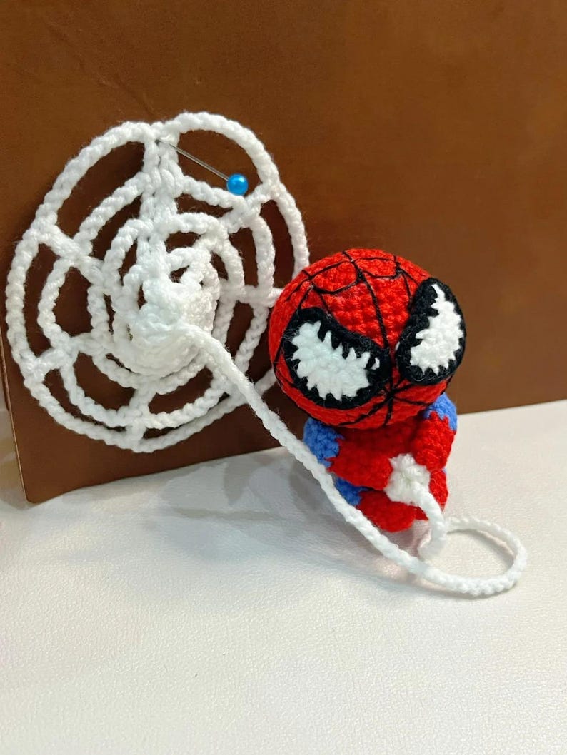 May include: A small, round, crocheted figure featuring a red and blue body with a large red head, black web-like stitching, and white eyes. The figure holds a long, white, crocheted spider web strand that connects to a circular white web piece.