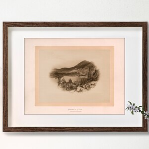 Può includere: Una stampa in bianco e nero incorniciata di una scena di lago di montagna. La stampa è in una cornice marrone con un passe-partout bianco. Il titolo della stampa è "Moosehead Lake".