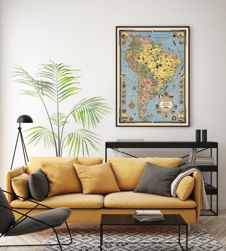May include: A vintage-style map of South America in a black frame hanging on a white wall above a yellow sofa. The map is filled with illustrations of animals, cities, and landmarks. The map is surrounded by a decorative border.