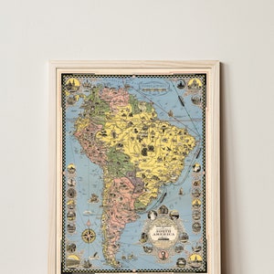 May include: A vintage illustrated map of South America with a blue background and yellow, green, and pink landmasses. The map is framed in a light wood frame. The map includes a scale of miles and a compass rose.