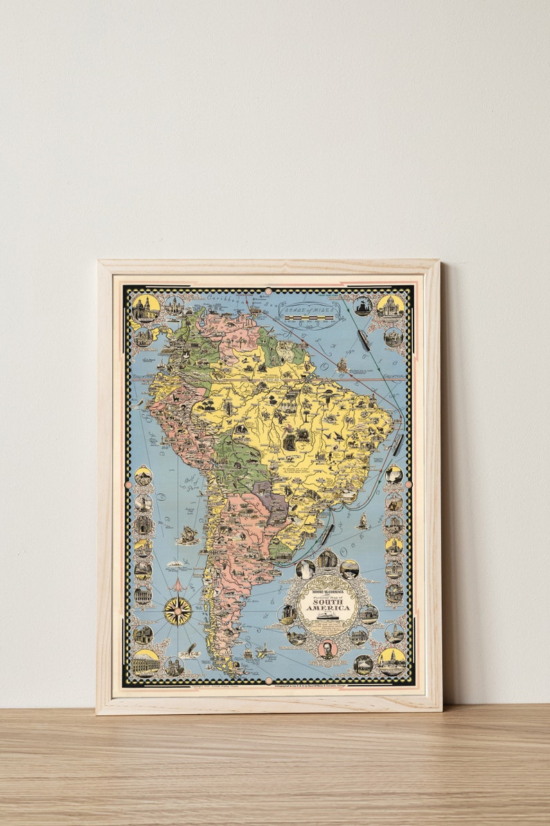 May include: A vintage illustrated map of South America with a blue background and yellow, green, and pink landmasses. The map is framed in a light wood frame. The map includes a scale of miles and a compass rose.