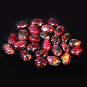 May include: A collection of oval-shaped red opals with iridescent flashes of color. The opals are arranged in a pile on a black background.
