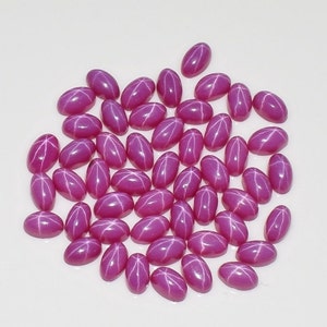 May include: A collection of oval-shaped, deep pink gemstones with a star-like pattern. The gems are scattered across a white surface, showcasing their polished surfaces and unique optical effect. These gemstones are ideal for jewelry making.